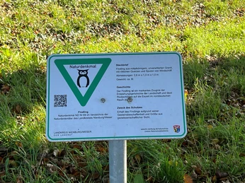 Findling Ein großer Findling dient als Naturdenkmal auf einer Wiese, flankiert von einem Informationsschild.