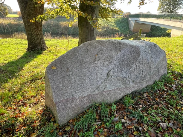 A large gray boulder in a meadow, shaded by trees, near a bridge over a river.