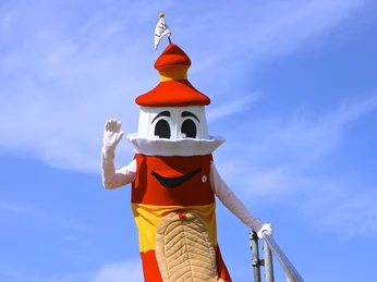 Lükko Leuchtturm vom Lükko Leuchtturm Kinderhaus Greetsiel Lükko Leuchtturm, das rot-gelbe Maskottchen, winkt lächelnd vor blauem Himmel auf einer Treppe