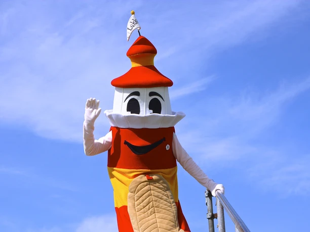 Lükko Leuchtturm vom Lükko Leuchtturm Kinderhaus Greetsiel Lükko lighthouse, the red and yellow mascot, smiling and waving against a blue sky on a staircase