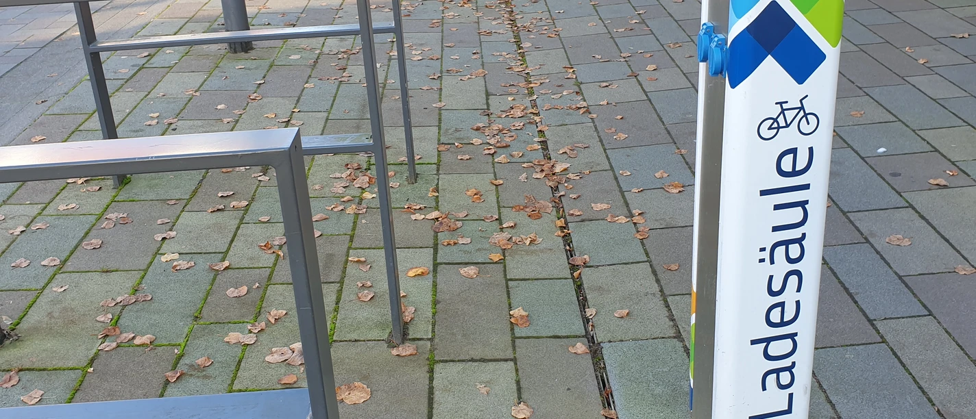 20241024_153701.jpg Eine E-Bike-Ladestation auf einem gepflasterten Gehweg, umgeben von Fahrradständern und Herbstlaub.