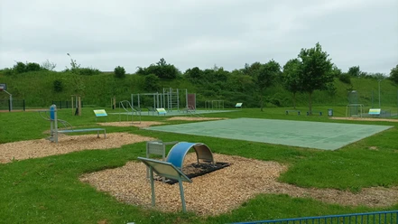 Outdoor-Fitnessbereich mit Geräten auf Rasenfläche, umgeben von Bäumen, mit Matten und Kiesflächen.