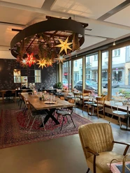 Innenraum des Cafés. Es steht ein großer Tisch in der Mitte auf einem Teppich. Rundherum stehen kleinere Tische an den Wänden, wovon eine Wand eine Fensterfront zur Straße hin ist. Von der Decke hängen leuchtende Papiersterne herab-
