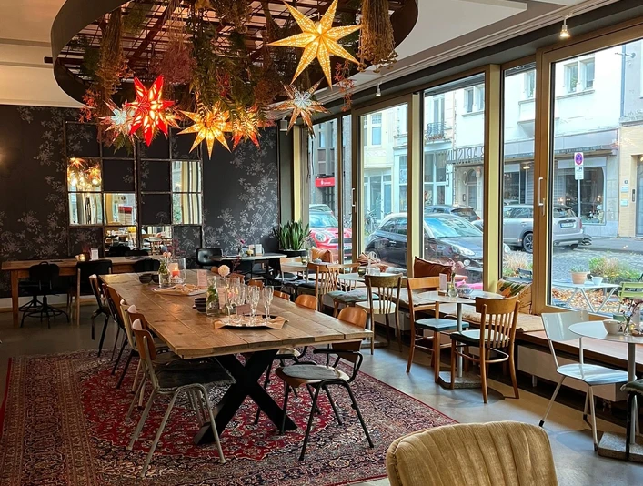 Café Innen Baristinho Innenraum des Cafés. Es steht ein großer Tisch in der Mitte auf einem Teppich. Rundherum stehen kleinere Tische an den Wänden, wovon eine Wand eine Fensterfront zur Straße hin ist. Von der Decke hängen leuchtende Papiersterne herab-