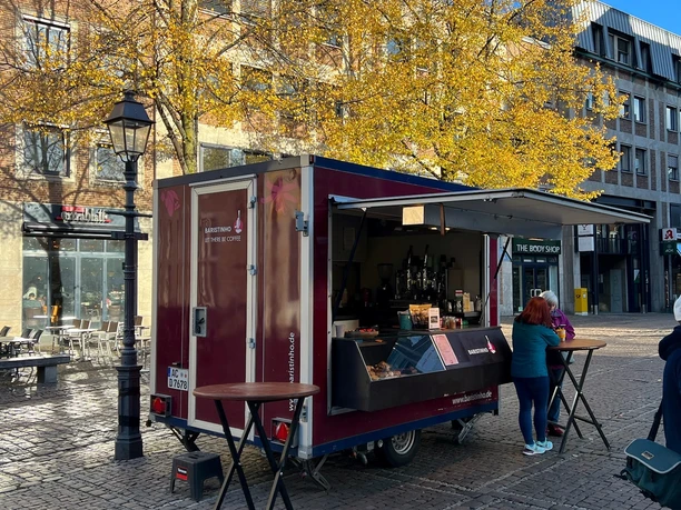 Vor einem Gebäude und einem großen herbstlichen Baum steht steht der Café-Wagen von Baristinho, der einseitig geöffnet ist und als Verkaufsstand genutzt wird. Davor stehen zwei runde Stehtische, an einem halten sich zwei Kundinnen auf.