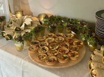 Catering Baristinho (c) Baristinho.jpg Auf einem vn einem Tischtuch bedeckten Tisch stehen verschiedene herzhafte Snacks, darunter Törtchen, in Gläsern geschichteter Salt und Wraps. Links brennen zwei Kerzen, rechts sind Teller gestapelt