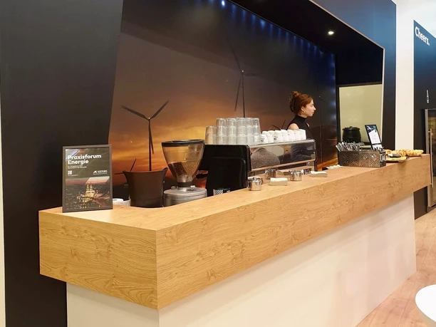 Catering-Stand mit hölzerner Theke. Hinter und auf der Theke stehen Gläser, Tassen, Besteckkörbe und eine Kaffeemaschine. Eine Frau steht hinter der Theke.