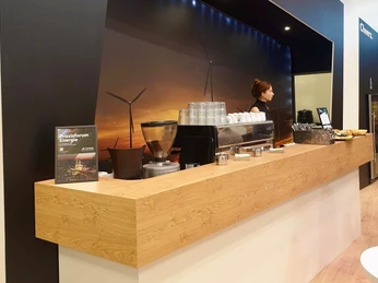 Catering-Stand Baristinho Catering-Stand mit hölzerner Theke. Hinter und auf der Theke stehen Gläser, Tassen, Besteckkörbe und eine Kaffeemaschine. Eine Frau steht hinter der Theke.