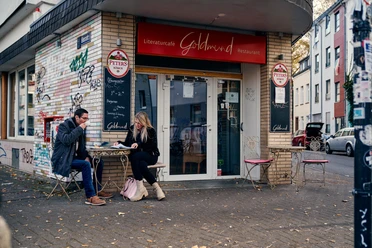 Goldmund Vor dem Literaturcafé Goldmund sitzen zwei Menschen an einem Tisch, umgeben von urbaner Graffiti-Ästhetik.