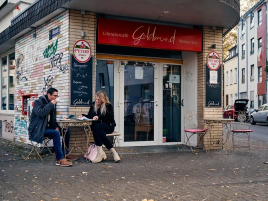Goldmund Vor dem Literaturcafé Goldmund sitzen zwei Menschen an einem Tisch, umgeben von urbaner Graffiti-Ästhetik.In front of the Literaturcafé Goldmund, two people are seated at a table, surrounded by an urban graffiti aesthetic.