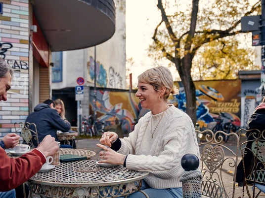 Goldmund Auf dem Bild sind mehrere Menschen zu sehen, die entspannt an einem runden Tisch im Freien sitzen. Sie genießen Getränke und unterhalten sich in einer freundlich gestalteten, urbanen Umgebung. Im Hintergrund sind eine kunstvoll bemalte Wand und herbstliche Bäume zu erkennen.The image shows several people comfortably seated around a round outdoor table, relaxing and enjoying drinks while engaging in conversation. They are situated in an inviting, urban environment. In the background, an artistically painted wall and trees bearing autumn colors can be seen.
