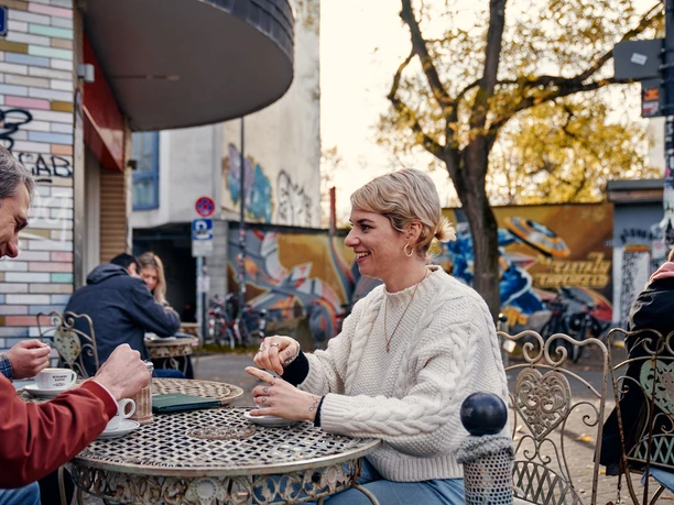 Goldmund Auf dem Bild sind mehrere Menschen zu sehen, die entspannt an einem runden Tisch im Freien sitzen. Sie genießen Getränke und unterhalten sich in einer freundlich gestalteten, urbanen Umgebung. Im Hintergrund sind eine kunstvoll bemalte Wand und herbstliche Bäume zu erkennen.