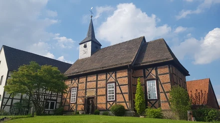 Ein Fachwerkgebäude mit Kirchturm und Wetterfahne vor blauem Himmel und flankiert von Bäumen.
