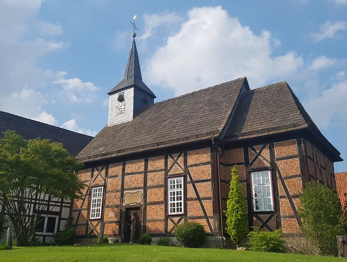 20210514_171537.jpg Ein Fachwerkgebäude mit Kirchturm und Wetterfahne vor blauem Himmel und flankiert von Bäumen.