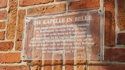 Transparente Plakette an einer Backsteinmauer erklärt die historische Bedeutung der Kapelle in Belle.