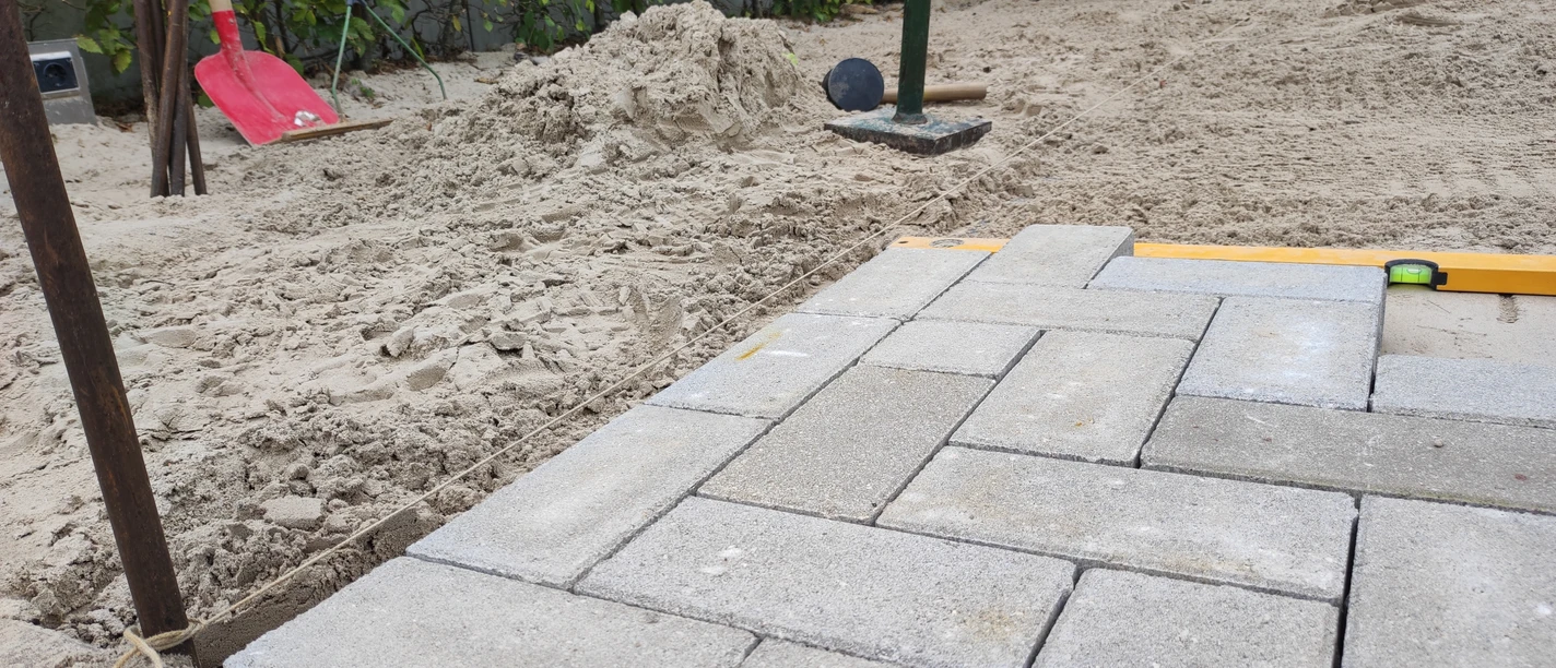 KiWiMedia_Webcode_23562360_Nadja Krause_2024-10-29.jpg Verlegen von Betonpflastersteinen auf Sandbett, Werkzeuge und Wasserwaage liegen daneben.