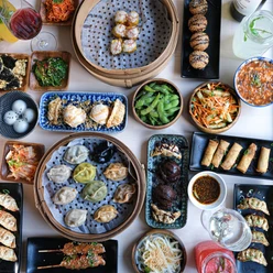 Tisch mit verschiedenen Asiatischen Spezialitäten wie Frühlingsrollen und Dumplings in unterschiedlichen Schalen, Tellern, Tabletts, Körben und Schüsseln.