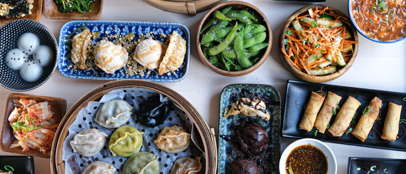 WeiundWei.jpg Tisch mit verschiedenen Asiatischen Spezialitäten wie Frühlingsrollen und Dumplings in unterschiedlichen Schalen, Tellern, Tabletts, Körben und Schüsseln.