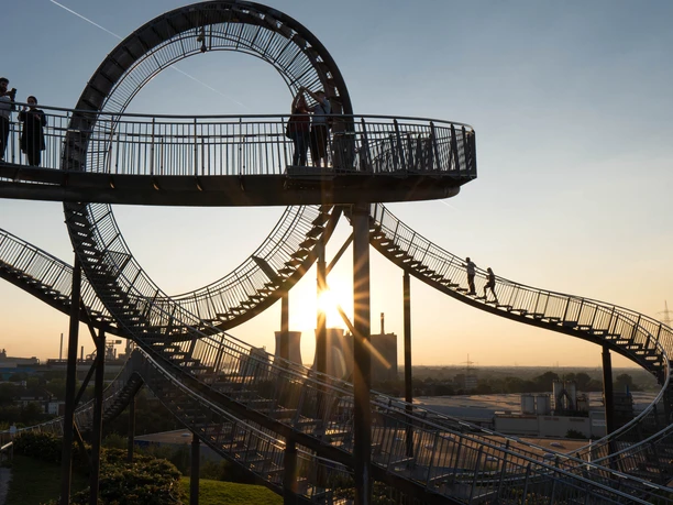 Tiger and Turtle Magic Mountain, Duisburg <p>Menschen spazieren auf der beeindruckenden Tiger & Turtle-Achterbahn-Skulptur bei Sonnenuntergang.</p>