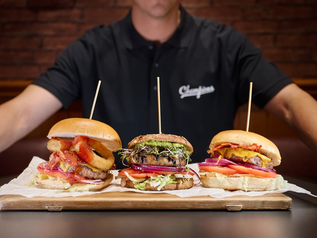 Champions Sports Bar - Gastronomie in Leipzig Ein Koch hält ein Holzbrett, auf dem drei Burger liegenA chef holds a wooden board with three burgers on itKuchař drží dřevěné prkénko se třemi hamburgery.Szef kuchni trzyma drewnianą deskę, na której leżą trzy burgeryEen chef-kok houdt een houten plank vast met drie hamburgers eropUno chef tiene in mano una tavola di legno con tre hamburger