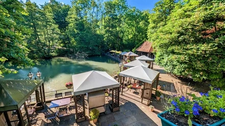 Der Terrassenbereich eines Hotels am Waldrand bietet Blick auf einen kleinen, von Bäumen umgebenen Teich.