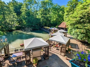 Der Terrassenbereich eines Hotels am Waldrand bietet Blick auf einen kleinen, von Bäumen umgebenen Teich.