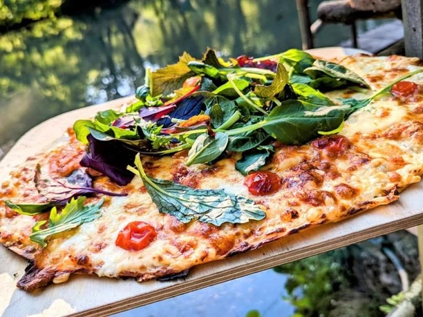 Bild13_©Waldhotel Silbermühle GmbH.jpg Flammkuchen auf Holzbrett, überdeckt mit frischen Kräutern, im Freien nahe eines ruhigen Gewässers.