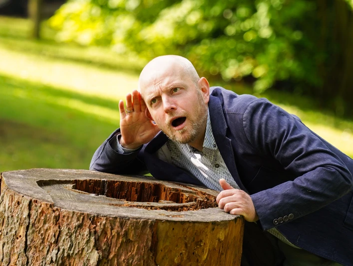 Familienkonzert_Ungarn_Fotocredit NWD_03.jpg Ein Mann lauscht aufmerksam an einem ausgehöhlten Baumstumpf im Grünen eines Parks.