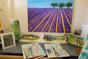 Auswahl Motive Andrea Moliere Mehrere Gemälde auf einem Tisch, darunter ein großes Bild eines violetten Lavendelfeldes mit Bäumen und kleinere Landschaftsmotive mit Felsen und Wäldern.