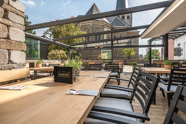Auszeit-Terrasse-2.jpg Überdachte Terrasse mit Holztischen und Stühlen, im Hintergrund eine alte Steinkirche bei Sonnenschein.