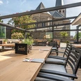 Auszeit-Terrasse-2.jpg Überdachte Terrasse mit Holztischen und Stühlen, im Hintergrund eine alte Steinkirche bei Sonnenschein.