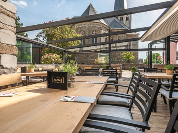 Überdachte Terrasse mit Holztischen und Stühlen, im Hintergrund eine alte Steinkirche bei Sonnenschein.