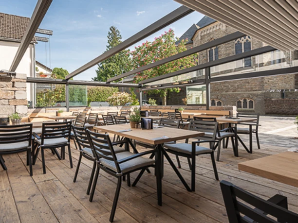 Ein sonnendurchfluteter Außenbereich eines Restaurants mit Holzboden, modernen Tischen und Stühlen.