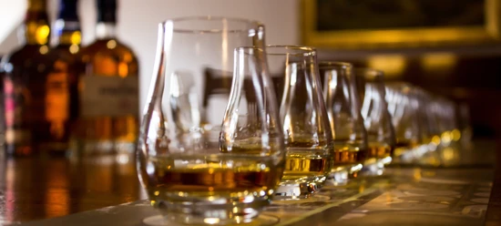 Eine Reihe von Whiskeygläsern auf einem Tisch, im Hintergrund unscharfe Whiskeyflaschen.