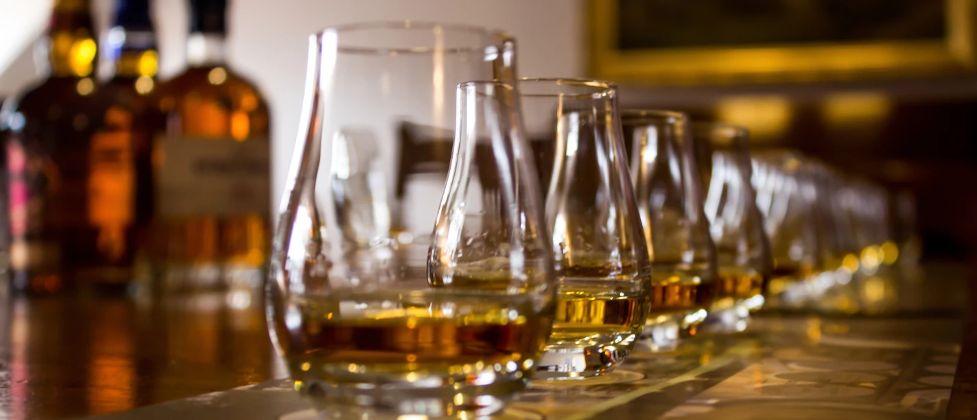Eine Reihe von Whiskeygläsern auf einem Tisch, im Hintergrund unscharfe Whiskeyflaschen.
