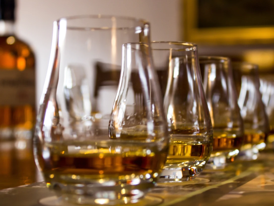 Bier- und Whiskey-Tasting.jpg Eine Reihe von Whiskeygläsern auf einem Tisch, im Hintergrund unscharfe Whiskeyflaschen.