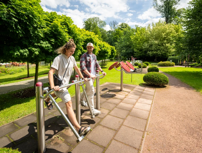 Zwei Personen nutzen Fitnessgeräte im Freien, umgeben von grünen Bäumen und einem gepflegten Weg im Kurpark.