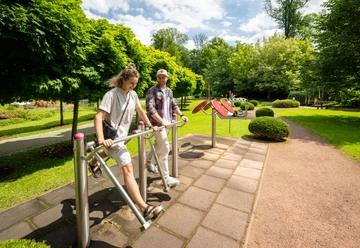 Kurpark Bad Holzhausen Zwei Personen nutzen Fitnessgeräte im Freien, umgeben von grünen Bäumen und einem gepflegten Weg im Kurpark.