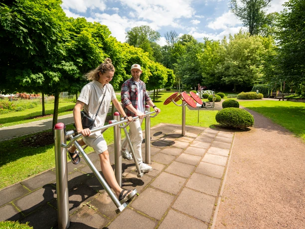 Kurpark Bad Holzhausen Zwei Personen nutzen Fitnessgeräte im Freien, umgeben von grünen Bäumen und einem gepflegten Weg im Kurpark.