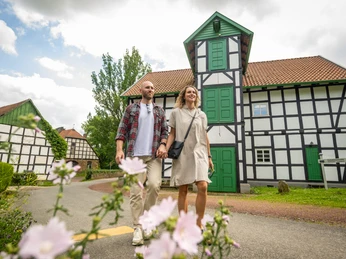 Ein Paar spaziert vor einer Fachwerk-Gutswassermühle, mit blühenden Blumen im Vordergrund.