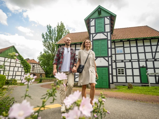 Ein Paar spaziert vor einer Fachwerk-Gutswassermühle, mit blühenden Blumen im Vordergrund.