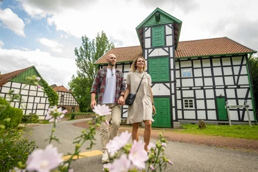Gutswassermühle Hudenbeck Ein Paar spaziert vor einer Fachwerk-Gutswassermühle, mit blühenden Blumen im Vordergrund.