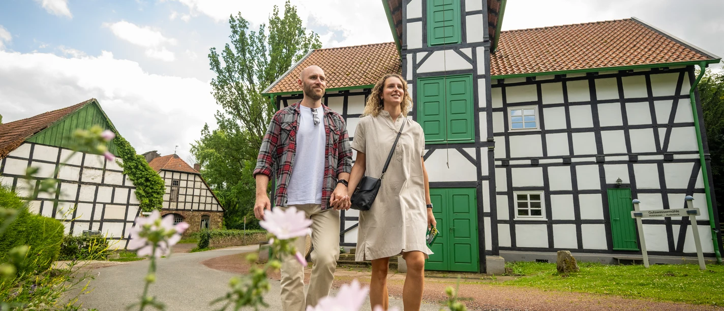Gutswassermühle Hudenbeck Ein Paar spaziert vor einer Fachwerk-Gutswassermühle, mit blühenden Blumen im Vordergrund.