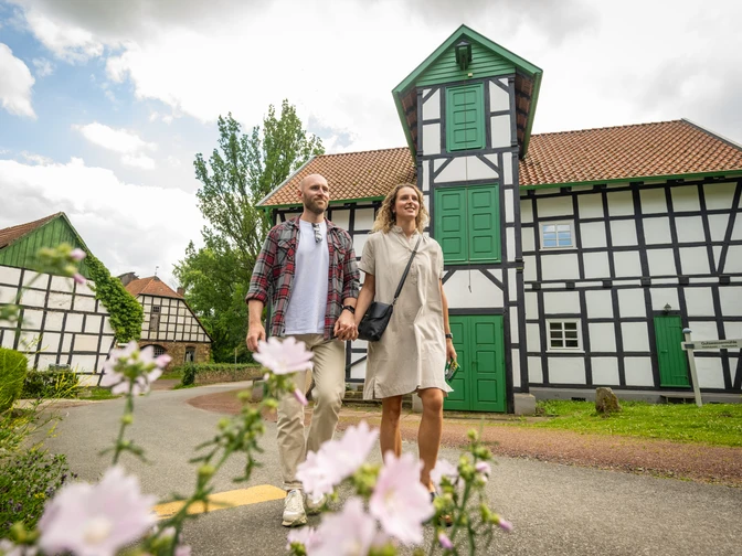 Gutswassermühle Hudenbeck Ein Paar spaziert vor einer Fachwerk-Gutswassermühle, mit blühenden Blumen im Vordergrund.