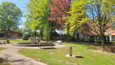 Offener Minigolfplatz mit Bäumen und Fachwerkhaus im Hintergrund, Wege und Sitzbänke vorhanden.