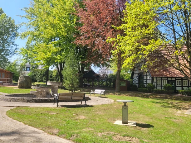 Offener Minigolfplatz mit Bäumen und Fachwerkhaus im Hintergrund, Wege und Sitzbänke vorhanden.