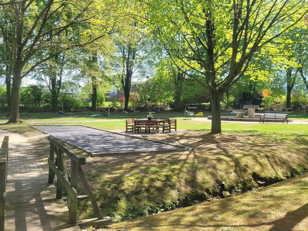 Grüner Minigolfplatz mit Holztischen, Bänken, Bäumen und einer kleinen Brücke im Sonnenlicht.
