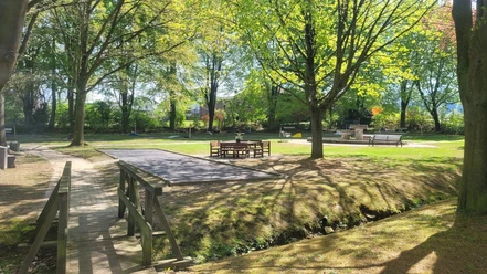 Minigolfplatz Börninghausen Grüner Minigolfplatz mit Holztischen, Bänken, Bäumen und einer kleinen Brücke im Sonnenlicht.