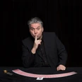 Nicht Blinzeln_BlackTableMagic.jpg Mann hält sich seinen Zeigefinder vor den Mund und stützt sich auf einen Tisch, auf dem Spielkarten halbkreisförmig ausgebreitet sind.