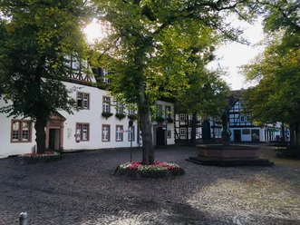 Der Kellerplatz in Beverungen zeigt ein weißes Rathaus mit Fachwerkgiebel. Ein Brunnen mit St.-Michaelsfigur steht im Mittelpunkt, umgeben von schattenspendenden Bäumen, die dem Platz eine einladende Atmosphäre verleihen. Diese Szene betont die historische und zugleich lebendige Präsenz des Ortes.